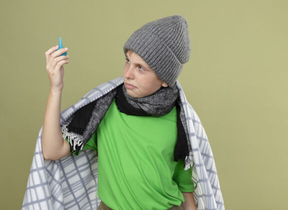 毯子生病的小男孩戴着暖和的帽子 围着围巾 裹着毯子 拿着灌肠剂 困惑而不高兴地站在轻墙上抱着困惑帽子