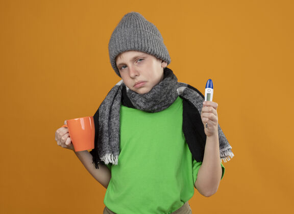 不舒服生病的小男孩穿着绿色t恤 戴着暖和的围巾 戴着帽子 手里拿着一杯热茶和温度计 站在橙色的墙上感到不舒服 不舒服和不开心感觉男孩杯子