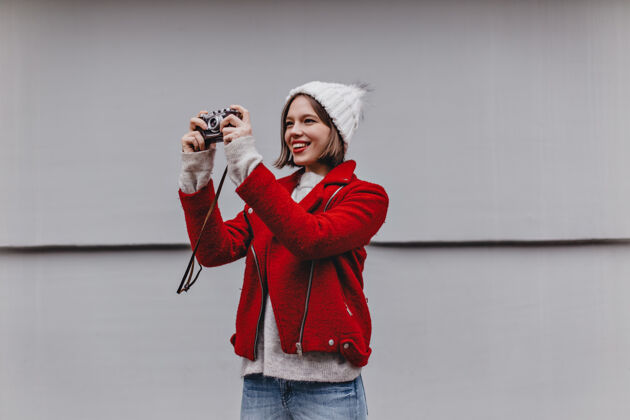 漂亮戴着针织帽子和红色外套的短发女人在复古相机上拍照人手套城市