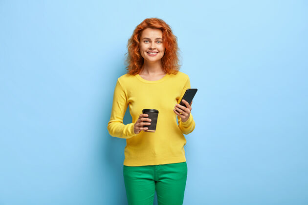 欢笑快乐的红头学生手里拿着现代手机 通过应用程序与朋友聊天通知毛衣一次性