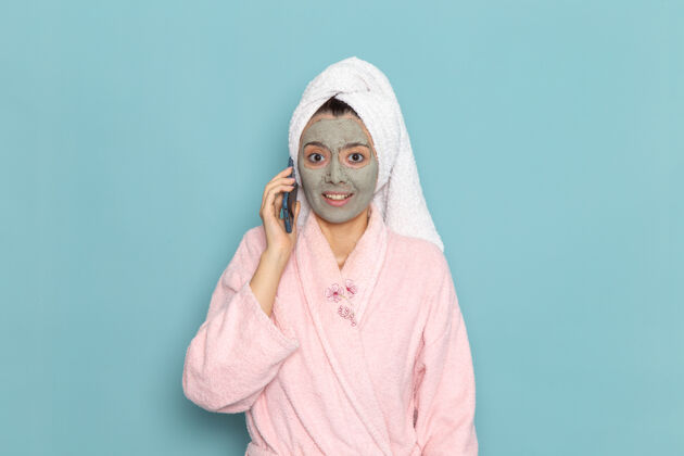 清洁正面图身着粉色浴袍的年轻女性淋浴后在蓝色墙壁上讲电话清洁美容净水自护霜淋浴视图肖像交谈