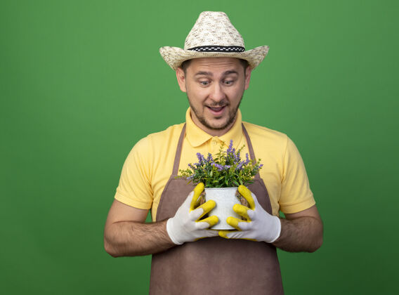 年轻人年轻的园丁穿着连体衣 戴着帽子 戴着工作手套 手里拿着盆栽植物 站在绿色的墙壁上惊讶地看着它植物抱工人