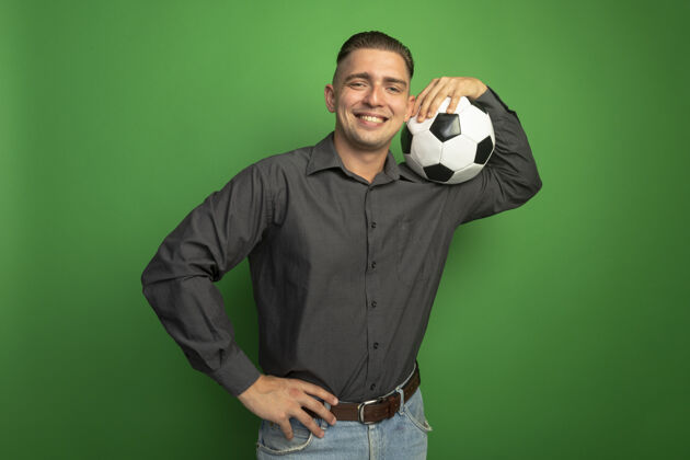 灰色身着灰色衬衫的年轻帅哥高兴地把足球扛在肩上 微笑着自信地站在绿色的墙上足球人衬衫