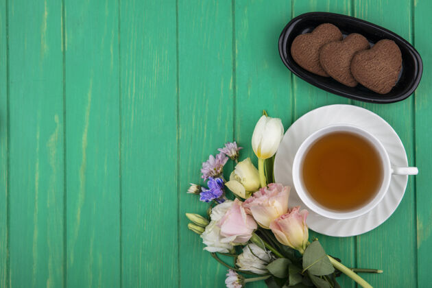 视图茶碟上的一杯茶和长方形碗中的心形饼干的俯视图 绿色背景上有鲜花和复印空间碗茶副本