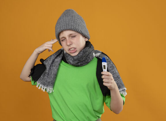 帽子生病的小男孩穿着绿色t恤 戴着暖和的围巾 戴着帽子 显示体温计 感觉不舒服 生病 不高兴 在站在橘色墙上的寺庙附近做着手势围巾体温计