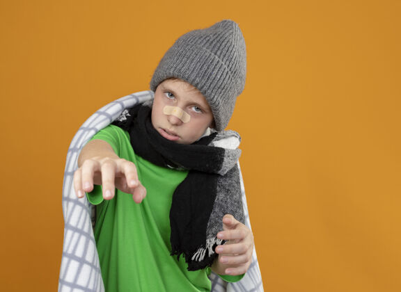 生病生病的小男孩戴着暖和的帽子 戴着围巾 裹着毯子 鼻子上贴着一块补丁 胳膊伸在橘色的墙上 看上去很不高兴 很不舒服橙色穿暖和