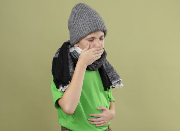 疾病生病的小男孩穿着绿色t恤 戴着暖和的围巾和帽子 站在光墙上咳嗽 感觉不舒服轻男孩帽子