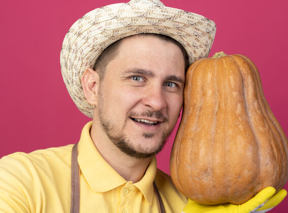 人年轻的园丁穿着连体衣 戴着帽子 戴着工作手套 手里拿着南瓜 站在粉红色的墙上 面带微笑地看着前面看男性微笑