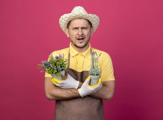 粉色年轻的园丁穿着连体衣 戴着帽子 戴着工作手套 手里拿着盆栽植物 站在粉红色的墙上 看着前面不高兴的样子男人站制服