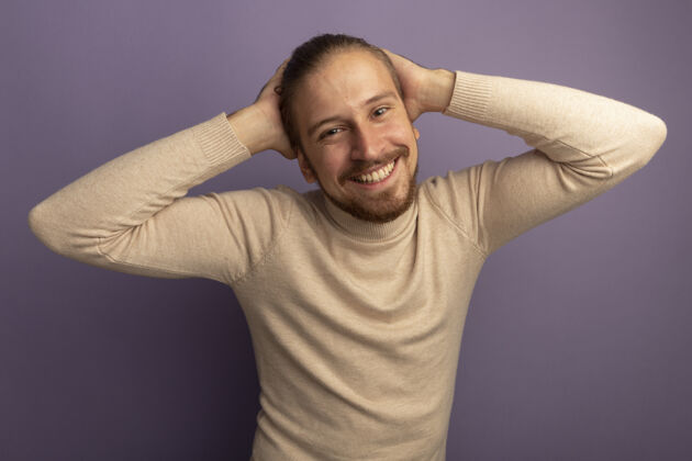 高领毛衣穿着米色套头衫的年轻帅哥站在淡紫色的墙上 面带微笑 双手放在头上人手丁香