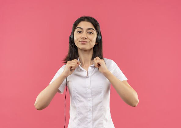 站立一个穿着白衬衫戴着耳机的年轻女孩 站在粉色的墙上摸着她的衣领 看起来很自信女性自信衬衫