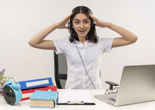 手势穿着白衬衫 戴着耳机的年轻女孩 双手抱着头 困惑地坐在桌子旁 把文件夹和笔记本电脑放在白墙上桌子人头