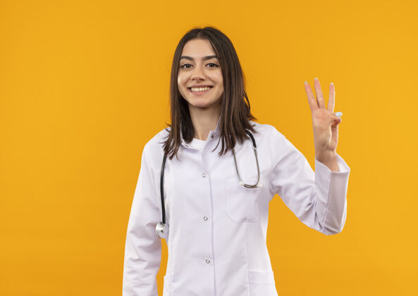 听诊器身穿白大褂的年轻女医生 脖子上戴着听诊器 手指指着三号站在橘色的墙上 脸上露出幸福的笑容女人外套三个