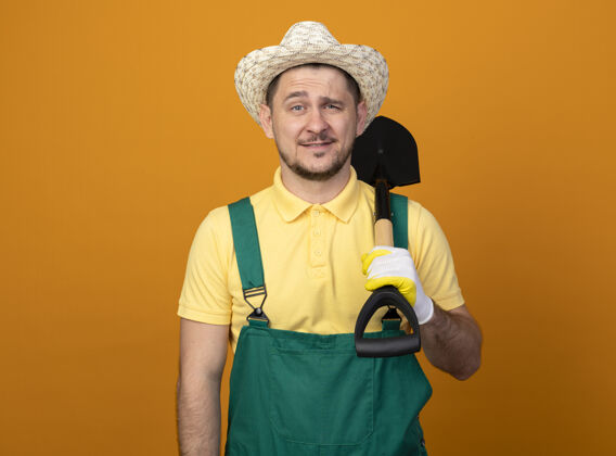 人年轻的园丁穿着连体衣 拿着铲子戴着帽子 站在橙色的墙上 微笑着自信地看着前方帽子微笑连身衣