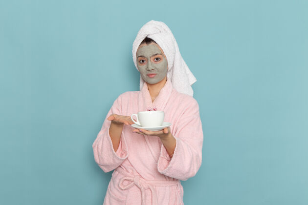 人正面图身着粉色浴袍的年轻女性手持一杯咖啡在蓝色墙壁上清洗美容自护霜淋浴清洁衣服咖啡