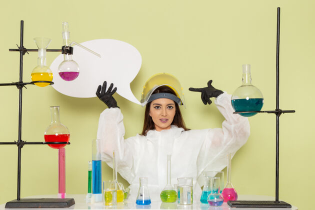 保持正面图身着特殊防护服的年轻女化学家手持绿色墙上的白色标牌工作化学实验室化学女科学保护实验室外套医学