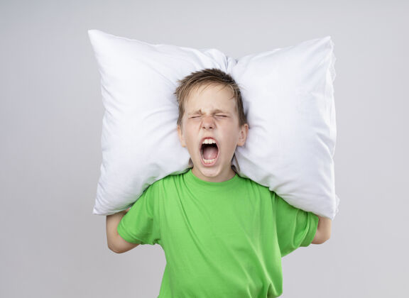 烦生病的小男孩穿着绿色t恤 站在白色的墙上 抱着枕头 带着恼怒的表情大喊大叫抱着喊小