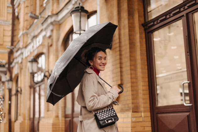 时尚身着米色风衣 背着十字形包 面带微笑的美女在欧洲城市的雨伞下行走亚洲微笑年轻人