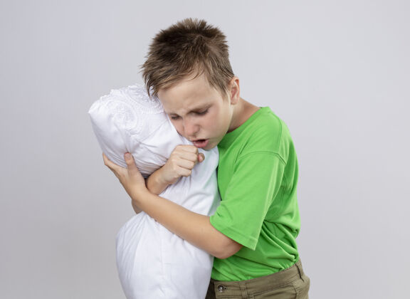 男孩生病的小男孩穿着绿色t恤感觉不舒服抱枕咳嗽站在白色的墙上枕头不舒服感觉