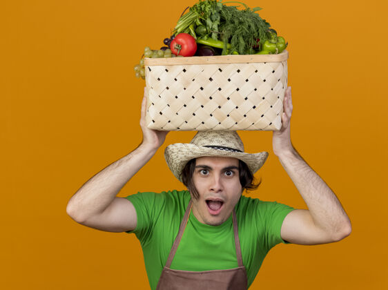年轻人年轻的园丁 围着围裙 戴着帽子 头上顶着装满蔬菜的箱子 站在橘色的墙上 满脸笑容板条箱围裙帽子