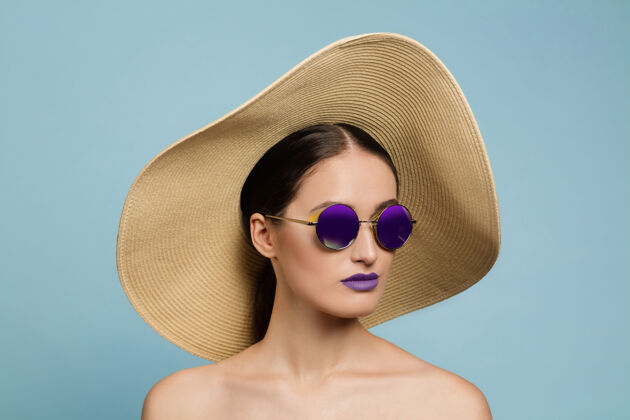 严肃美丽女人的肖像 明亮的妆容 蓝色工作室背景上的帽子和太阳镜时尚和时尚的制作和发型夏天的颜色美丽 时尚和广告概念严肃头发女性人