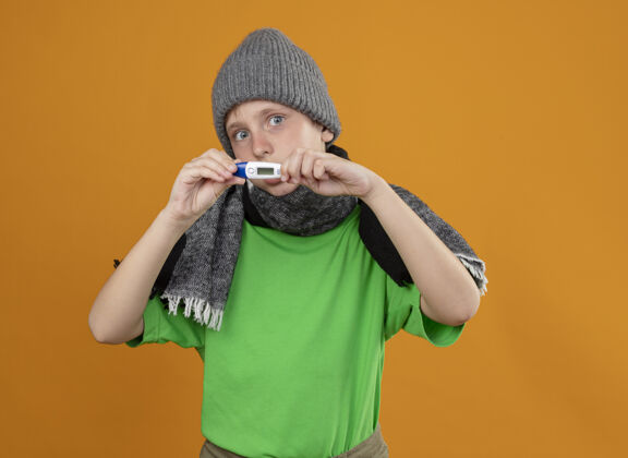 不开心生病的小男孩穿着绿色t恤 戴着暖和的围巾和帽子 站在橙色的墙上 显示体温计不舒服 生病 不开心生病不舒服感觉