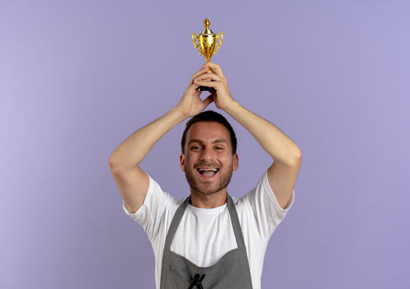 工人围裙里的理发师把奖杯举过头顶 站在紫色的墙上 高兴而兴奋男人T恤抱着