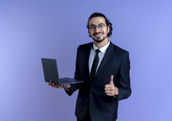 站着一位身穿黑色西装 戴着眼镜 手持笔记本电脑 面带微笑 竖起大拇指的商人站在蓝色的墙上公司市民优雅