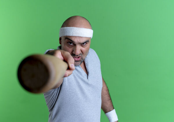 棒球戴着头巾的成熟的运动型男人拿着棒球棒指着前面 愤怒的脸站在绿色的墙上脸人小伙子