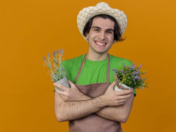 微笑年轻的园丁 围着围裙 戴着帽子 手里拿着盆栽植物 站在橙色的墙上 双手交叉 面带微笑站着手花园