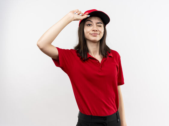 制服身穿红色制服 戴着帽子的年轻送货员站在白墙上 微笑着 眨着眼睛摸着帽子站摸微笑