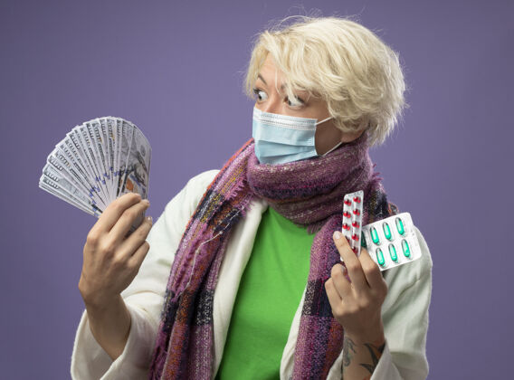 生病生病的不健康的女人 短发 戴着暖和的围巾 戴着护面面具 手里拿着现金和药丸 站在紫色的墙上 看起来很困惑 很担心 有疑虑看着药丸不健康
