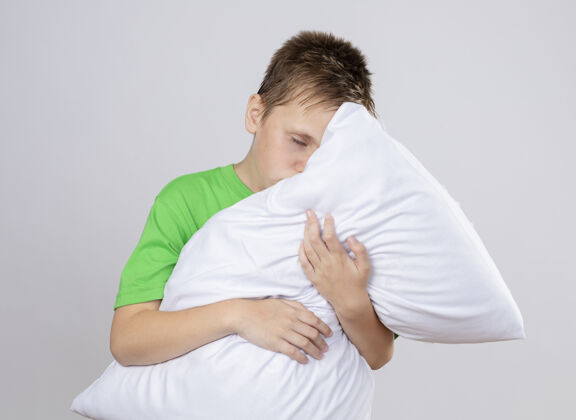 拥抱生病的小男孩穿着绿色t恤感觉不舒服抱枕闭着眼睛站在白色的墙上疾病枕头小