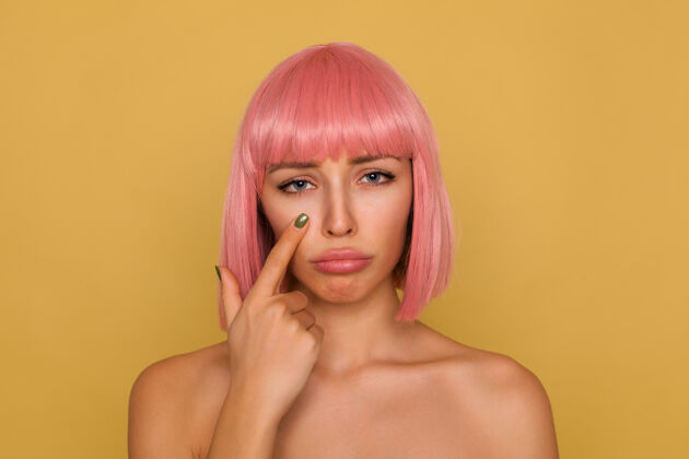发型不快乐的年轻迷人的粉红头发女性 鲍勃的发型撅着嘴唇 神情悲伤 站在芥末墙上时 食指夹在眼睛下面提高20岁食指