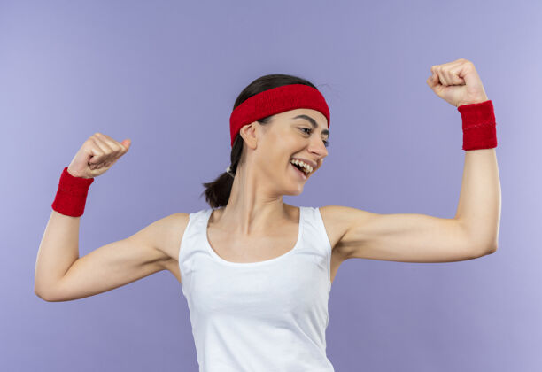 健康身穿运动服 头戴花边 微笑着 高举拳头的年轻健身女士站在紫色的墙上 快乐而积极拳头站立运动装