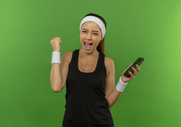 姿势身穿运动服 头戴智能手机的年轻健身女士高举拳头站在绿色的墙壁上 快乐而兴奋拳头人运动装