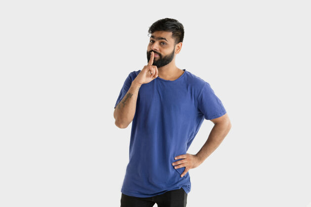 站立美丽的男性半身肖像隔离在白色的工作室背景年轻感性的印度教男子在蓝色衬衫面部表情 人类的情感 广告概念思考 窃窃私语的秘密消极年轻手势