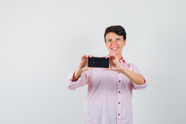 女人穿着粉色衬衫的女性在手机上拍照 看起来很愉快 正面视图快活年轻人服用