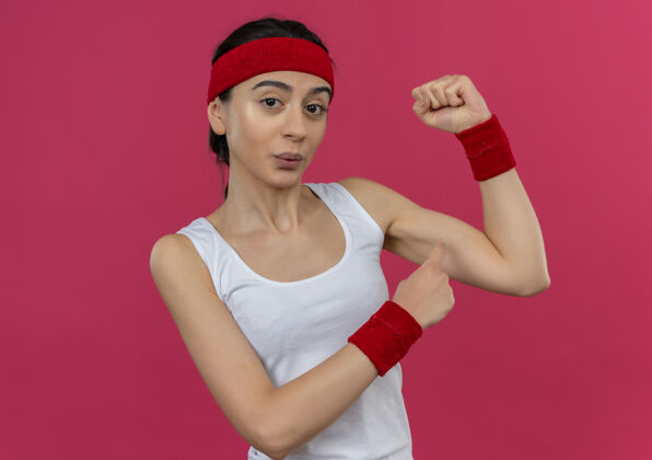 信心身着运动服的年轻健身女士戴着头巾举起拳头 站在粉红色的墙上 露出自信的二头肌年轻看拳