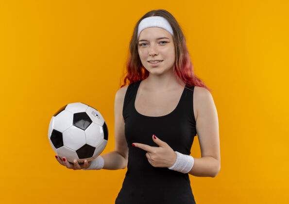 姿势身着运动服的年轻健身女士站在橙色的墙上 手里拿着足球 用食指指着它微笑着手指目录足球