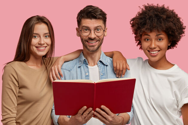 种族开朗混血的横拍三位友好的团友紧紧地站在一起 拿着红色的课本 为共同的任务阅读信息 隔着粉色的墙教育理念高加索人非洲伴侣
