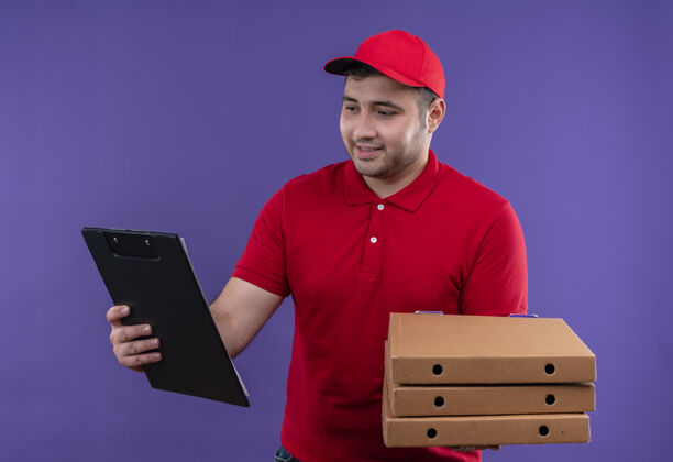 市民身穿红色制服 头戴鸭舌帽 手持披萨盒和剪贴板的年轻送货员站在紫色的墙上 脸上洋溢着幸福的笑容递送帽子站着