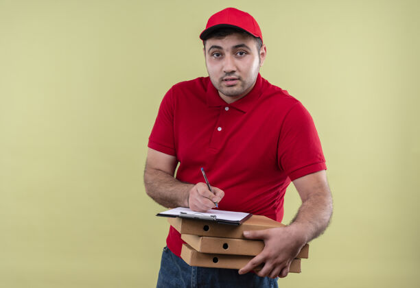 年轻人年轻的送货员身穿红色制服 戴着帽子 手里拿着披萨盒 站在绿色的墙上友好地微笑着写在空白页上书写站着姿势