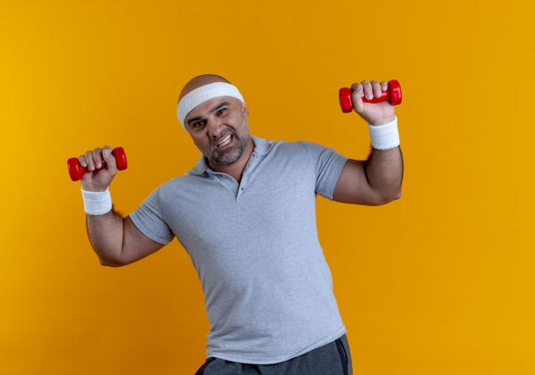 运动戴着头巾的成熟运动型男子站在橙色的墙上 举着看起来很紧张的哑铃锻炼身体紧张男人教练