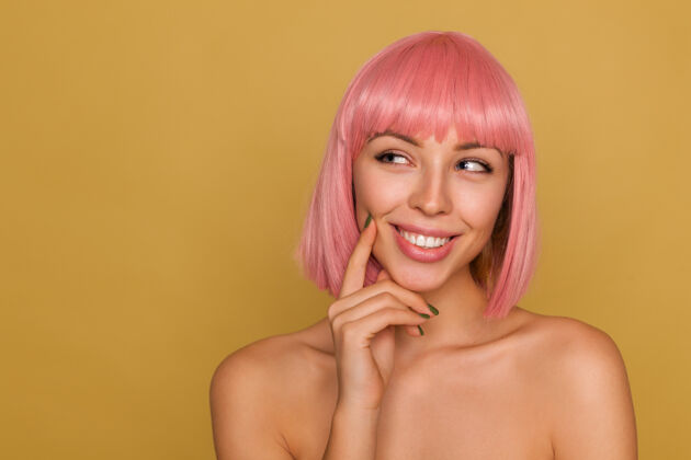 站立迷人沉思的年轻粉红头发女性 自然妆容 食指贴在脸颊上 站在芥末墙前 愉快地微笑着爱情手姿势