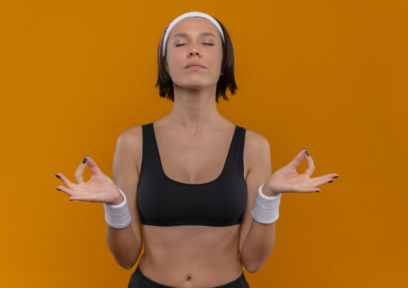 冥想穿着运动服 戴着头巾 闭着眼睛放松 手指站在橙色的墙上做冥想姿势的年轻健身女士运动运动女人