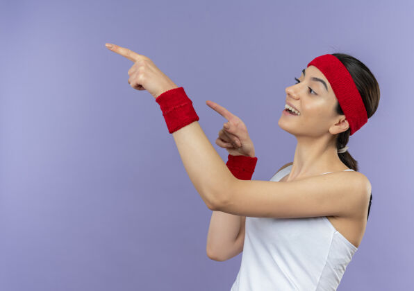 健身身穿运动服的年轻健身女士戴着头巾 一边惊讶地看着一边 一边用手指指着紫色的墙壁年轻旁边运动装