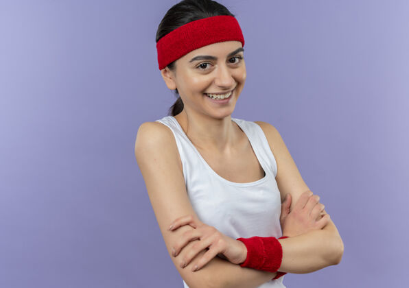 人身穿运动服 头戴花边 双臂交叉胸前 笑容满面的年轻健身女士站在紫色的墙上健身运动员手臂