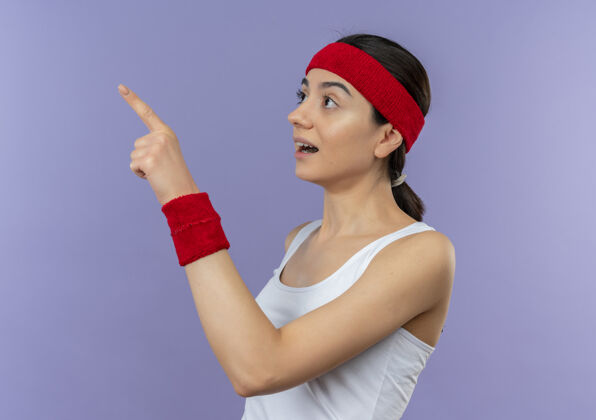 运动身穿运动服的年轻健身女士戴着头巾 一边惊讶地看着一边 一边用手指指着紫色的墙壁惊喜壁板健身