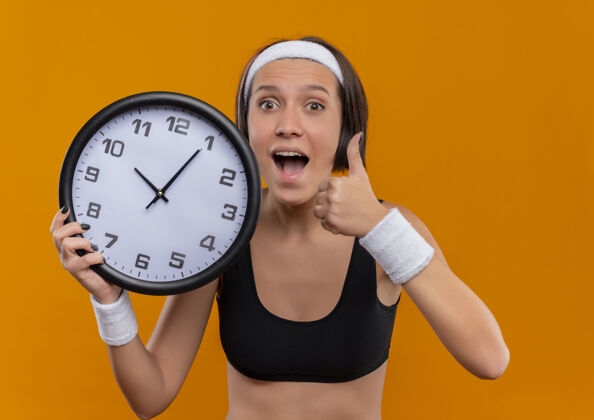 女人身穿运动服的年轻健身女士 头箍举着挂钟 高兴而兴奋地竖起大拇指站在橙色的墙上时钟女人站着